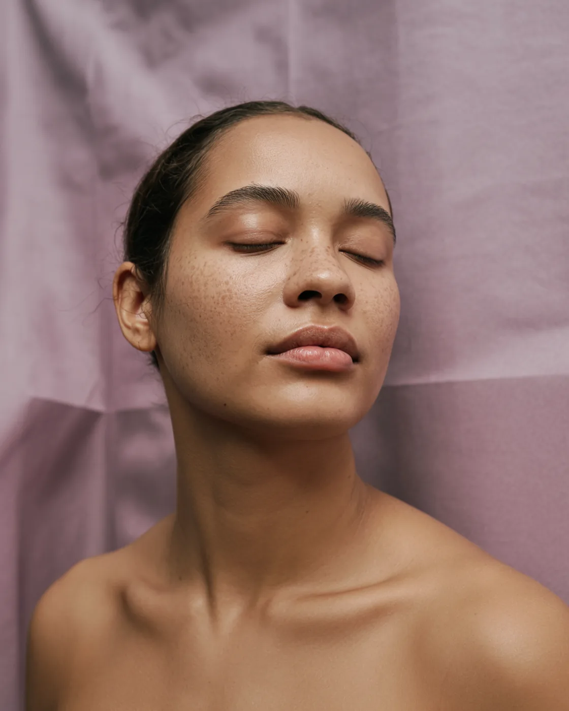 Mulher de 22 anos de olhos fechados, ombros nus, pele natural com sardas sobre fundo malva claro