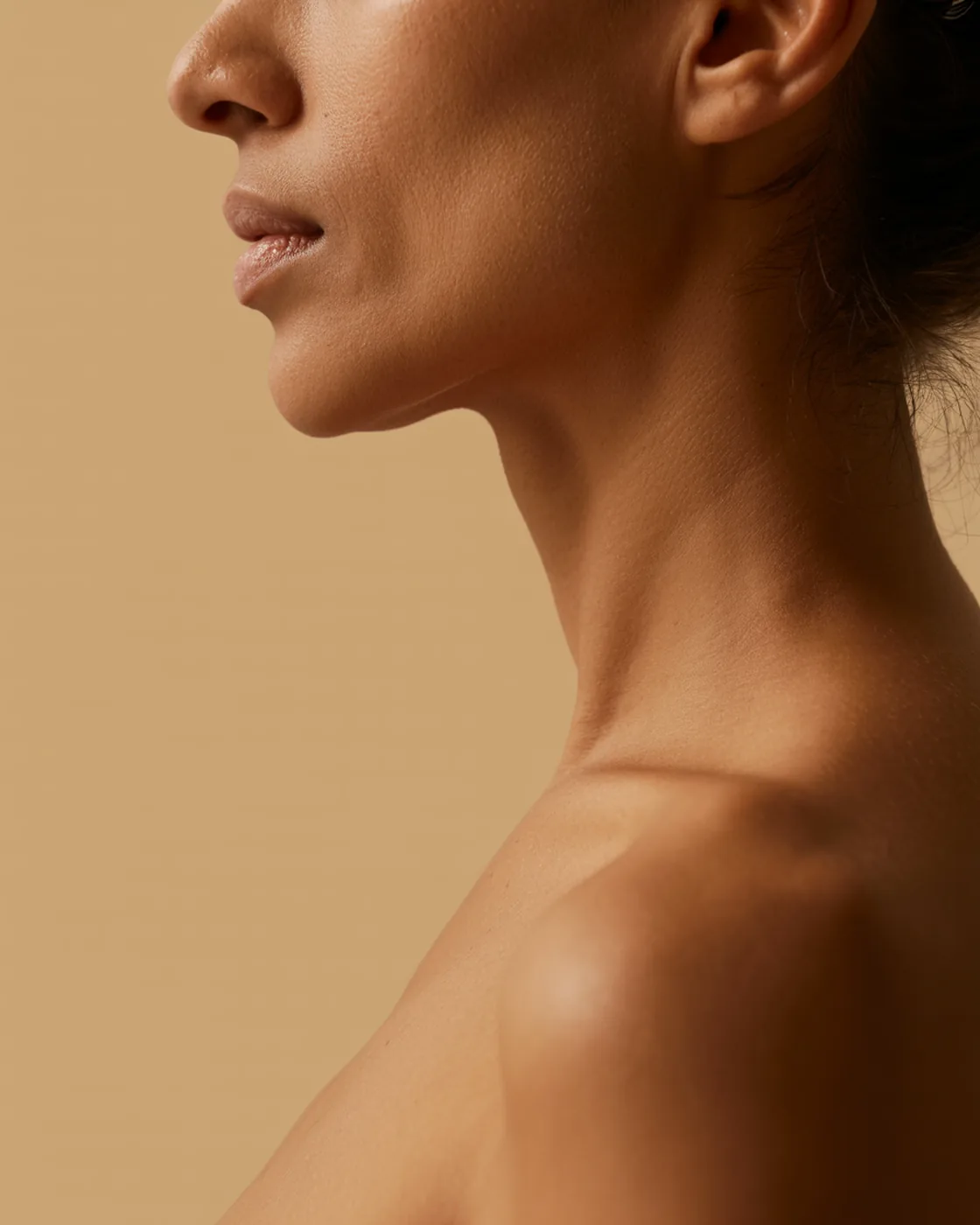Mulher de 35 anos de perfil, cortada do nariz para baixo, pescoço e ombro com pele natural sobre fundo aveia