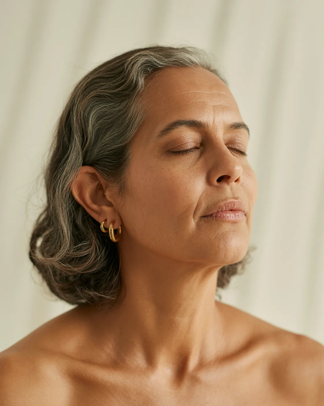 Mulher de 55 anos com cabelo grisalho natural e brincos de argola, olhos fechados, pele natural sobre fundo creme