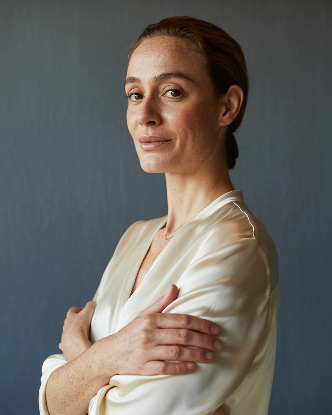 Retrato da dermatologista responsável pela clínica, de blusa de seda creme e cabelo preso, braços cruzados, olhando para a câmera sobre fundo cinza-escuro com luz de janela