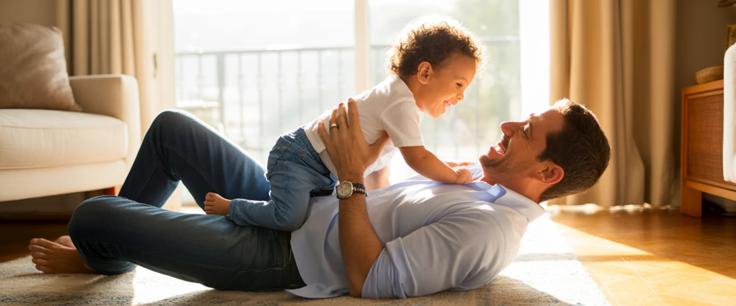 Pai deitado no tapete da sala segurando o filho pequeno no alto, os dois rindo, luz dourada de fim de tarde