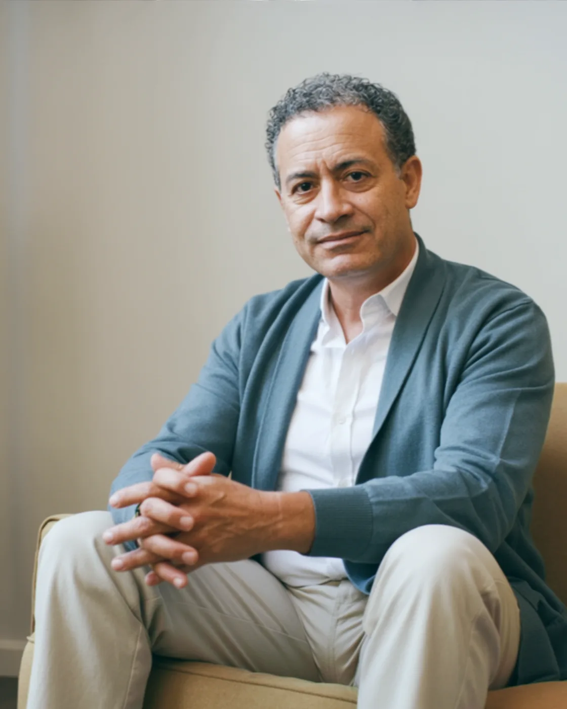 Dr. Tomás Ribeiro, psiquiatra, sentado com as mãos no colo, cardigã cinza-azulado, diante de uma parede clara em luz de janela