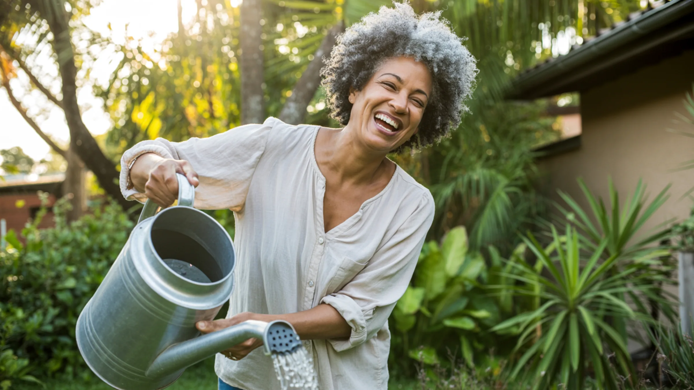 Mulher de cabelo grisalho rindo enquanto rega as plantas do jardim ao entardecer