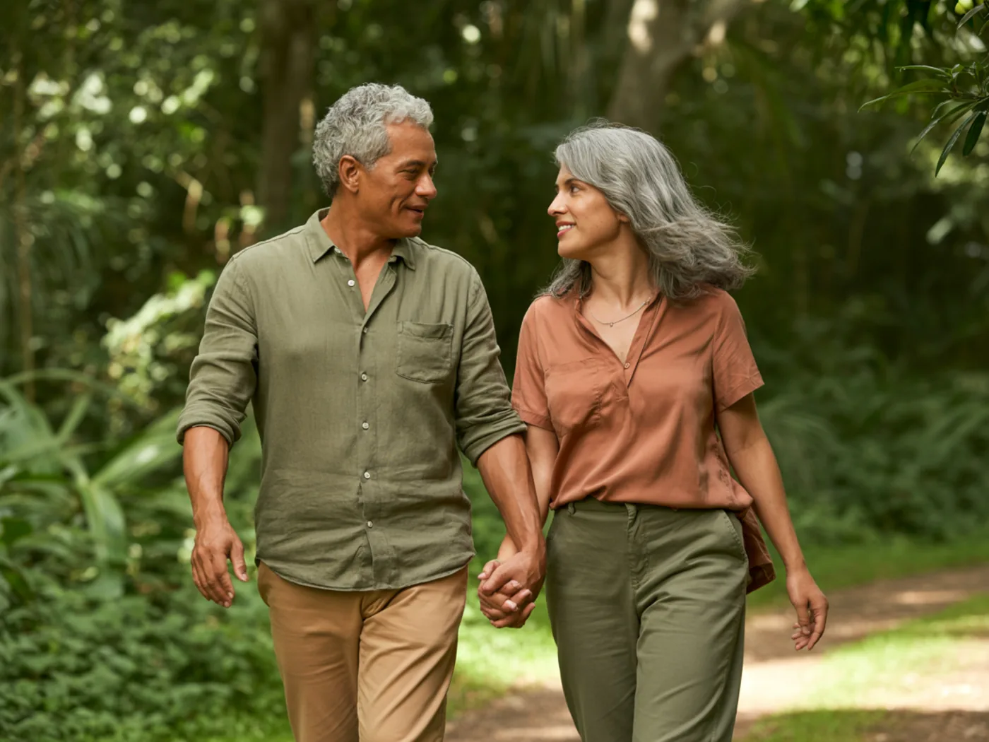 Casal de sessenta anos caminhando de mãos dadas por uma trilha de mata, com o sol entrando entre as árvores