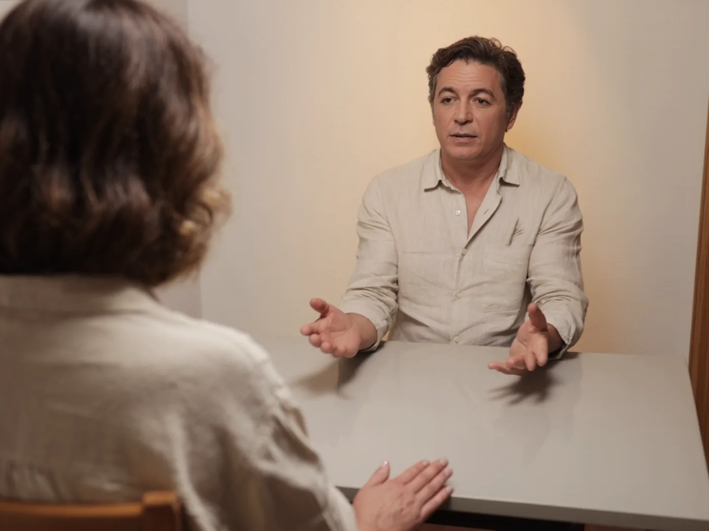 Cirurgião sentado à mesa branca do consultório explicando com as mãos abertas a uma paciente vista de costas e fora de foco